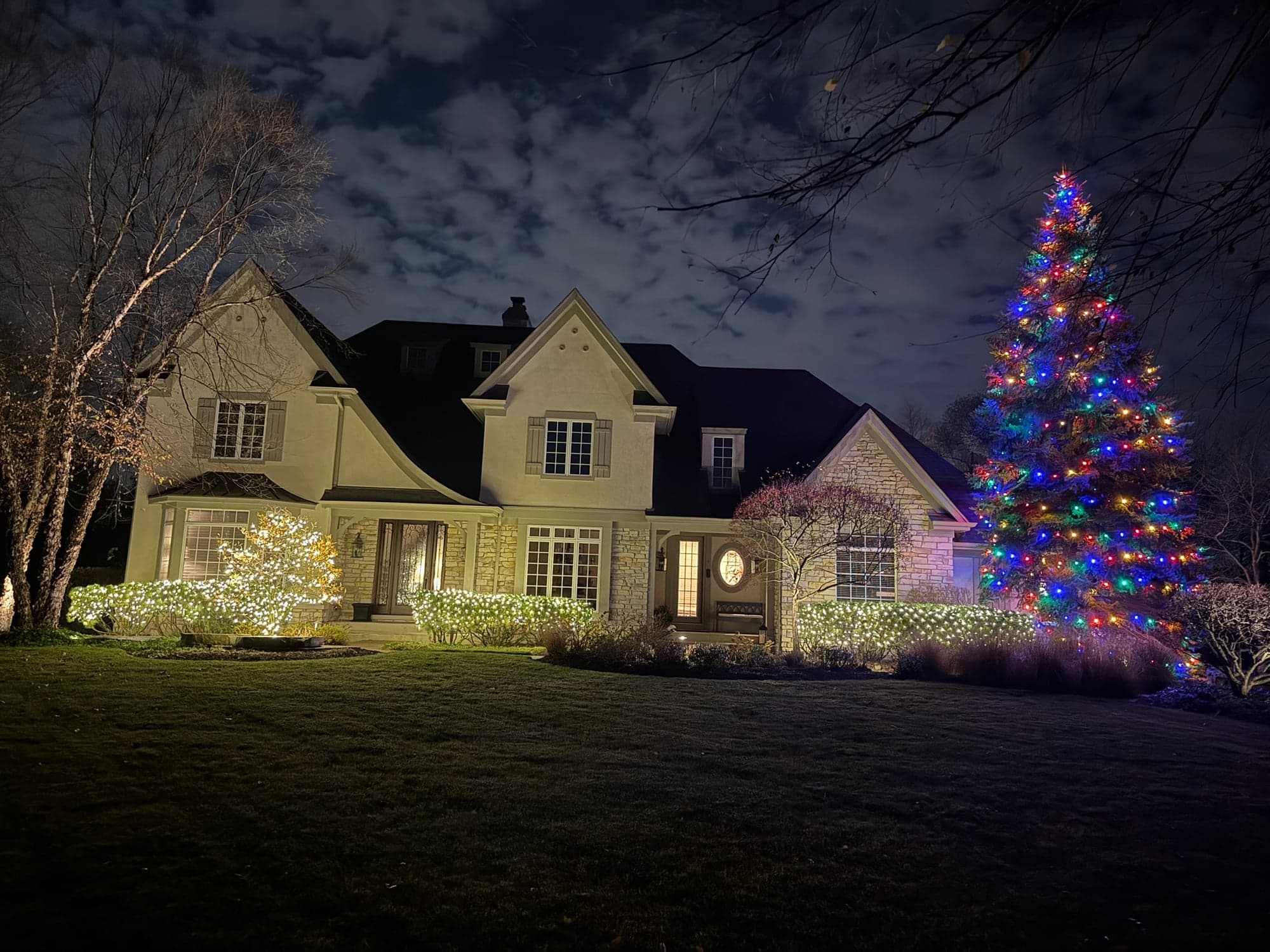 Stuccoed two-story home with a tall multicolor-lit Christmas tree and white-net-lit bushes along the front